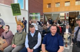 People sit in rows in a meeting. Two people holds signs that read "Fracking injects toxins into Mother Earth" and "Protect."