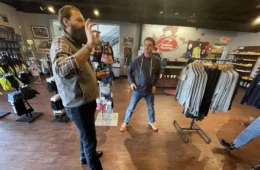 Two men stand inside a store with clothing and other outdoor gear.