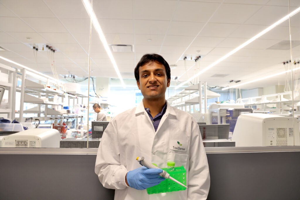 Aravind Krishnan is wearing a white lab coat standing in a la