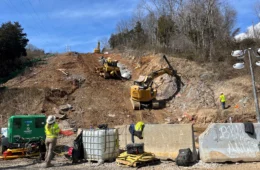 Construction equipment on a cleared hillside, with a worker in the foreground, behind concrete barriers