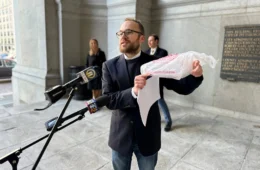 A man holds a plastic bag as he talks in a microphone outside a buidling