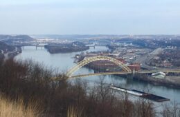 C barge full of coal travels up the Ohio River under a yellow bridge.