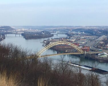 C barge full of coal travels up the Ohio River under a yellow bridge.