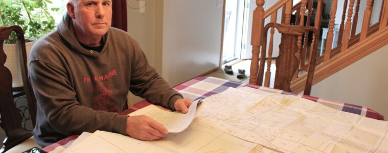 A man sits at a dining room table with a large document showing property lots