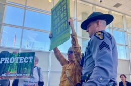 A woman stands holding a sign above her head with a state trooper standing next to her.
