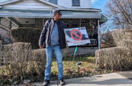 A woman stands in front of a house holding a sign that reads, "Stop SOBE."