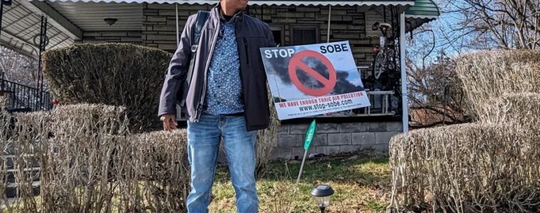 A woman stands in front of a house holding a sign that reads, "Stop SOBE."