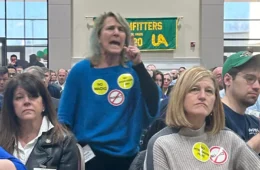 A woman in a blue shirt with protest pins stands in an audience yelling. In front of her are two ther women wearing pins.