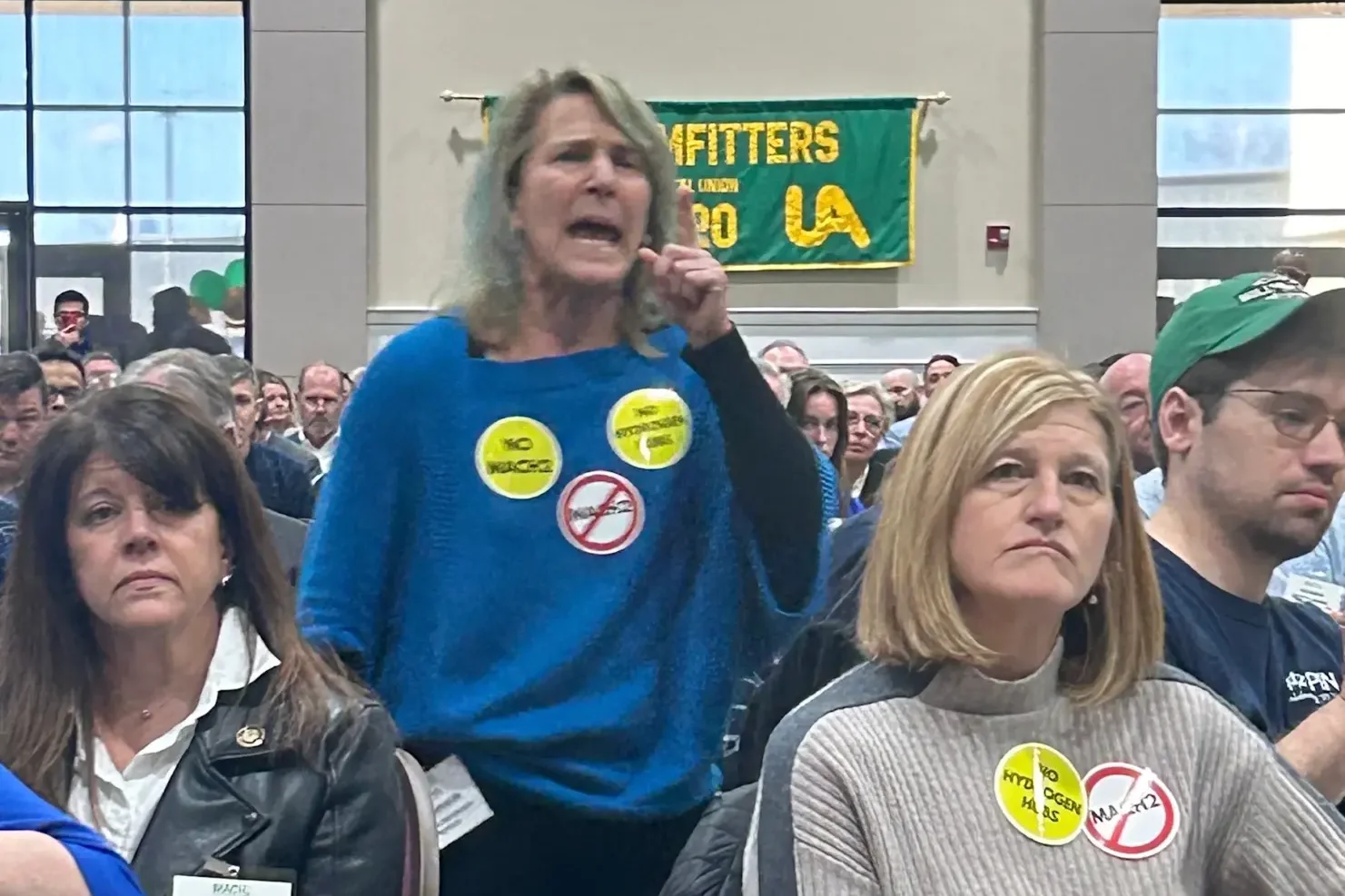 A woman in a blue shirt with protest pins stands in an audience yelling. In front of her are two ther women wearing pins.
