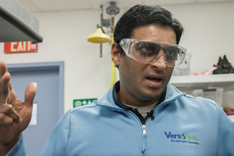 A man with safety goggles and a blue shirt stands in a lab.