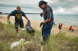 Two men using poles with grabbers on the ends pick up trash in the brush near the beach