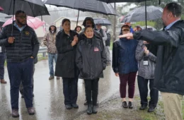 Deb Haaland in a black rain coat under an umbrella, along with a crowd of people under umbrellas in the rain.