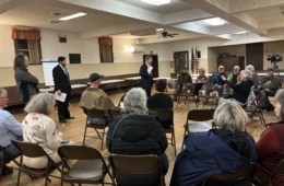 People sit in a circle on folding chairs listening to a man standing in the middle of the room.