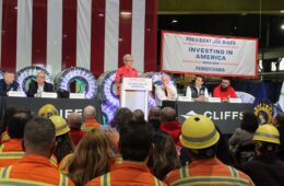 A woman stands at a podium with a row of men sitting at a table behind her, and a seated crowd in front, including workers in orange jackets and yellow hardhats.