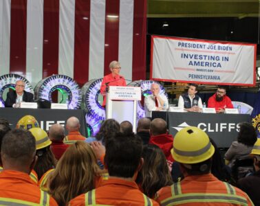 A woman stands at a podium with a row of men sitting at a table behind her, and a seated crowd in front, including workers in orange jackets and yellow hardhats.