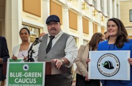 A man in a cap, tie and vest stands behind a podium with a green sign that reads "Blue-Green Caucus." A woman in a suit stands next to him holding a sign that reads "Pennsylvania House, Blue Green Caucus."