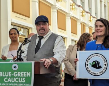 A man in a cap, tie and vest stands behind a podium with a green sign that reads "Blue-Green Caucus." A woman in a suit stands next to him holding a sign that reads "Pennsylvania House, Blue Green Caucus."