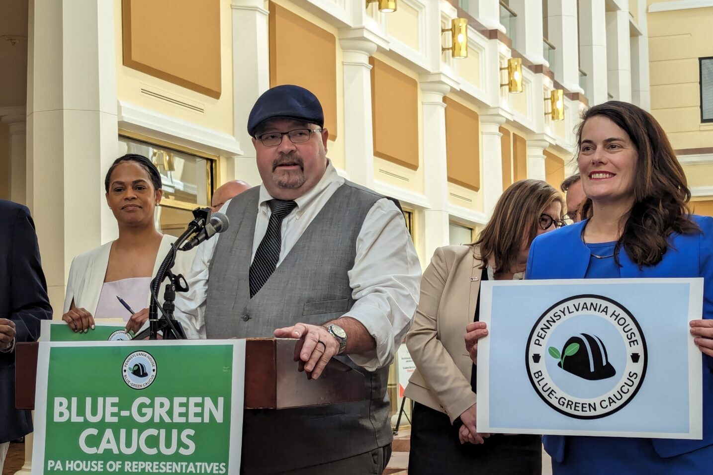 A man in a cap, tie and vest stands behind a podium with a green sign that reads "Blue-Green Caucus." A woman in a suit stands next to him holding a sign that reads "Pennsylvania House, Blue Green Caucus."