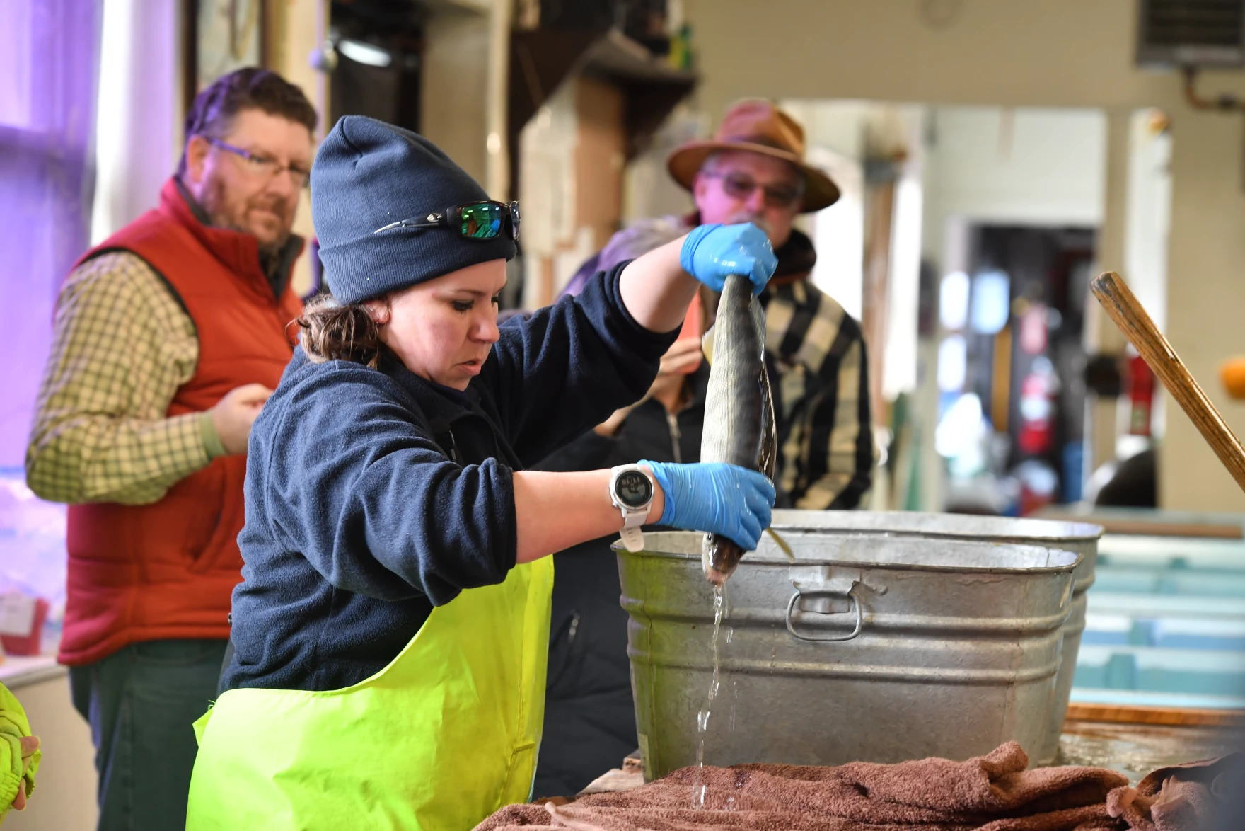 A woman in a knit cap and yellow waders holds a fish upside down as water drips off of it, as a crowd looks on.