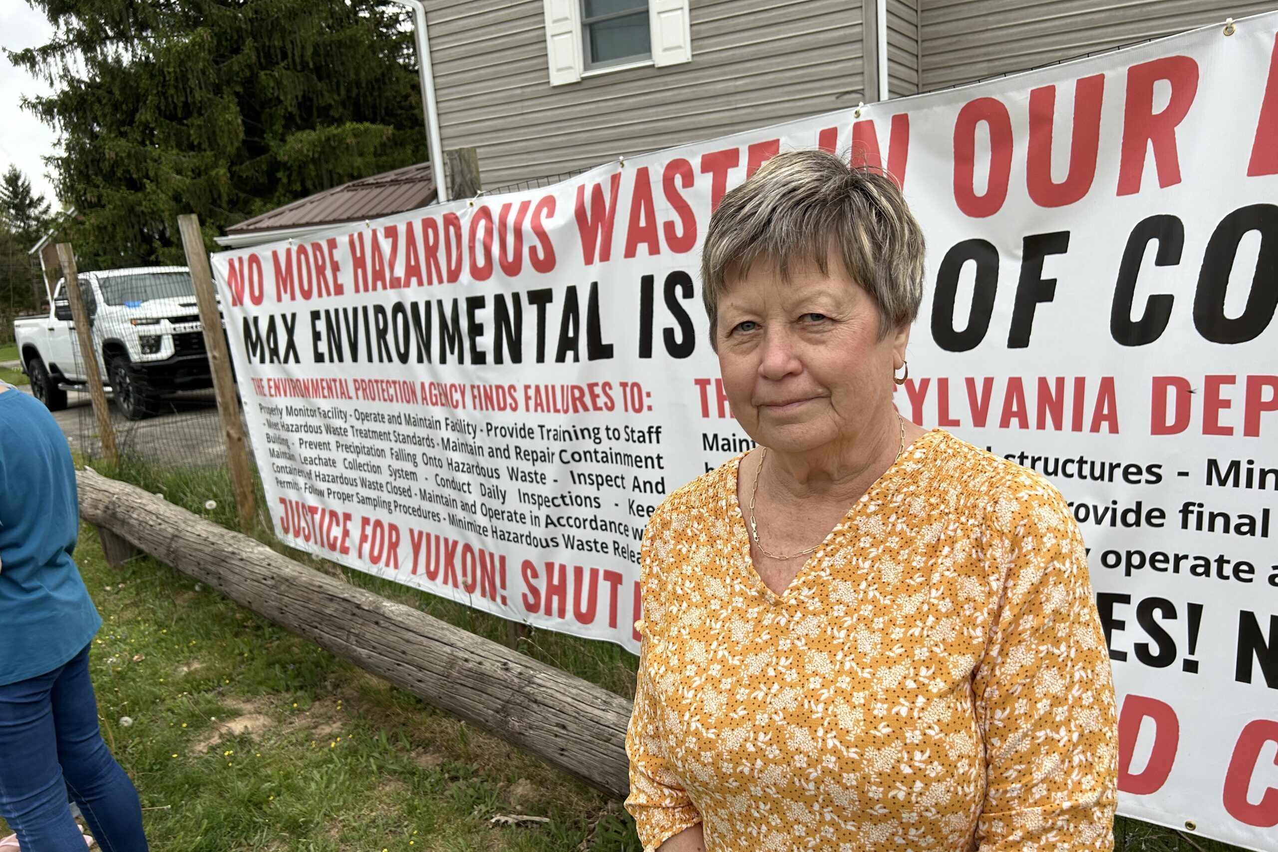 A woman in a yellow shirt stands next to a long sign on a fence that reads in part, "No More Hazardous Waste...Max Environmental"