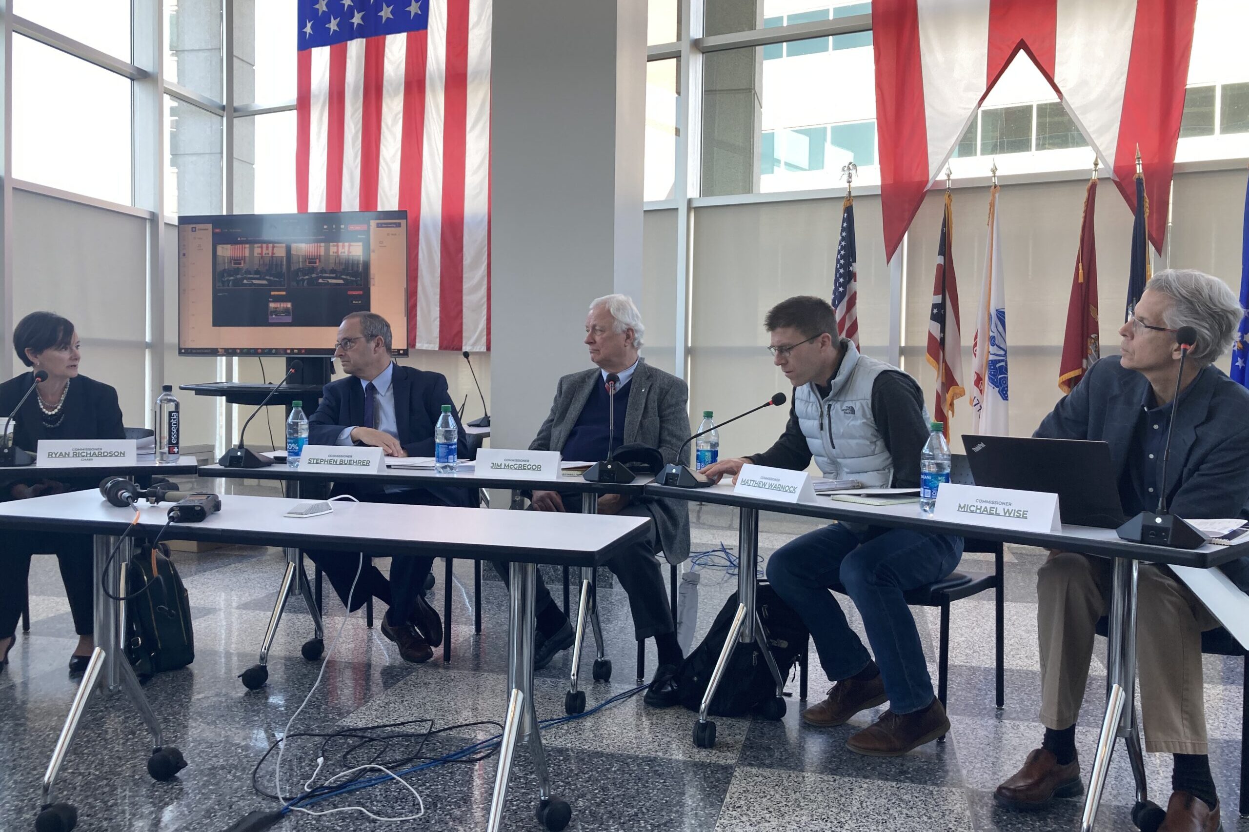 A panel of five people sit at tables in front of an American flag