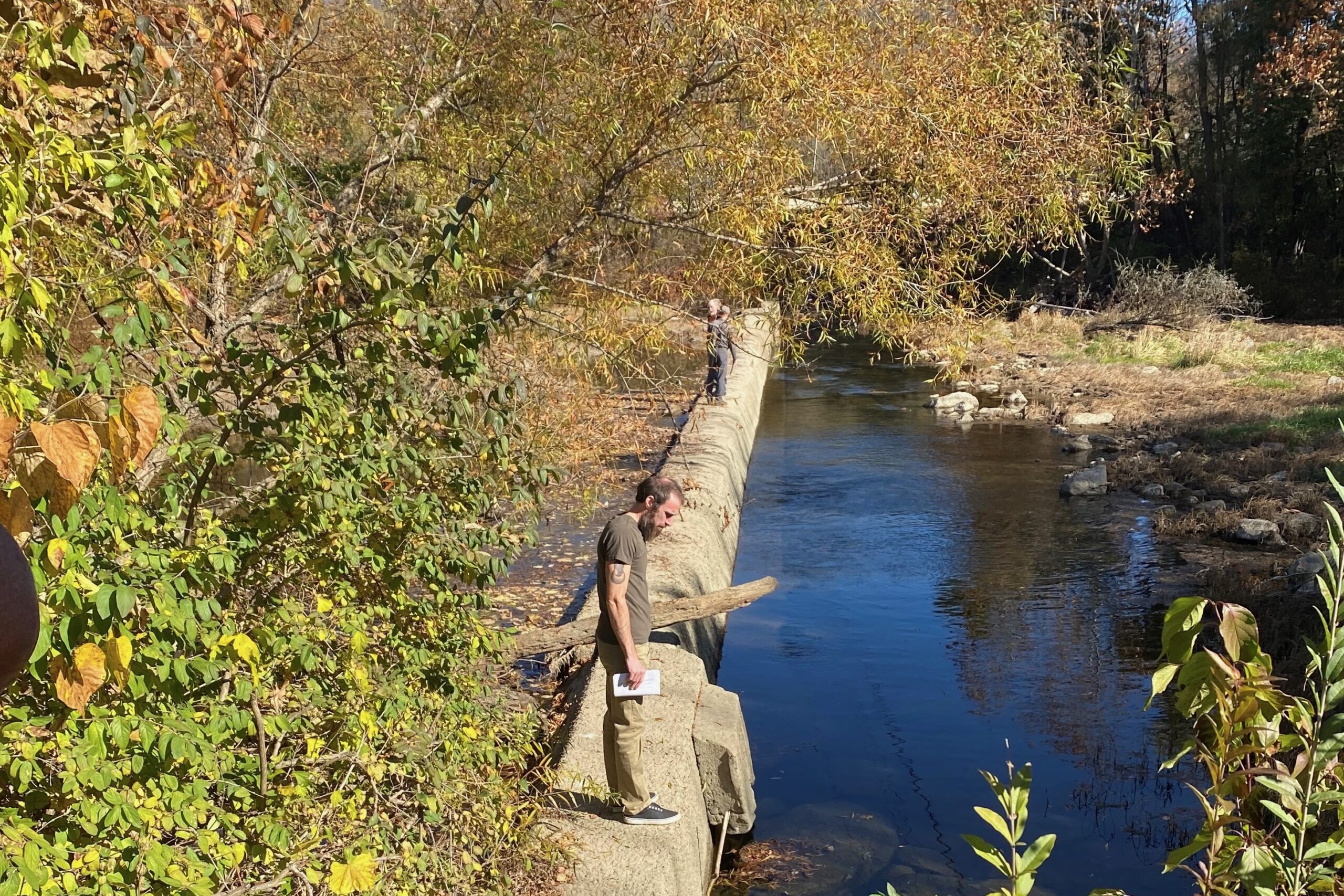 A man stands on a concrete barrier that crosses a wide stream.