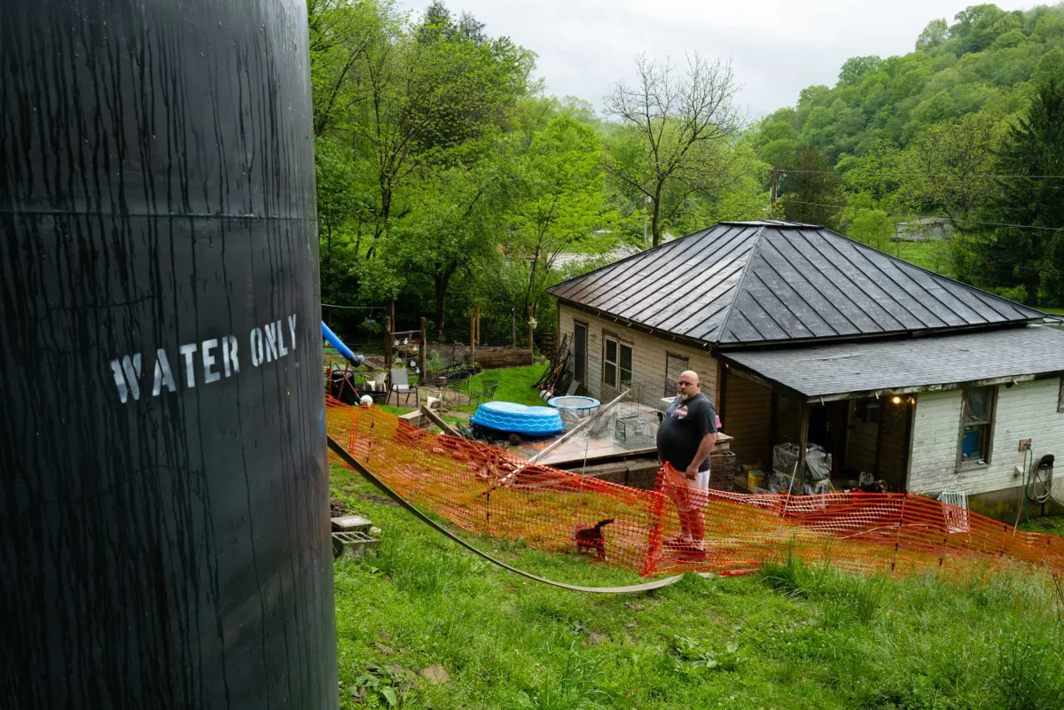 A man stands down hill from a black tank in the backyard of his home