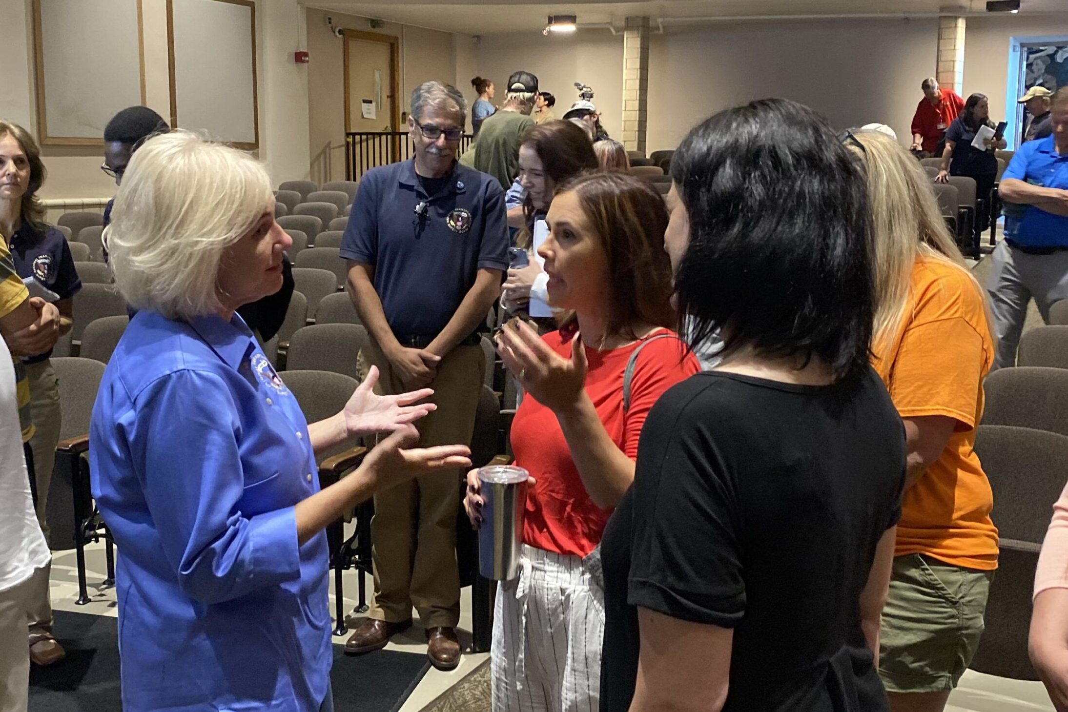 A woman in a blue shirt standing in a school auditorium talking with three women