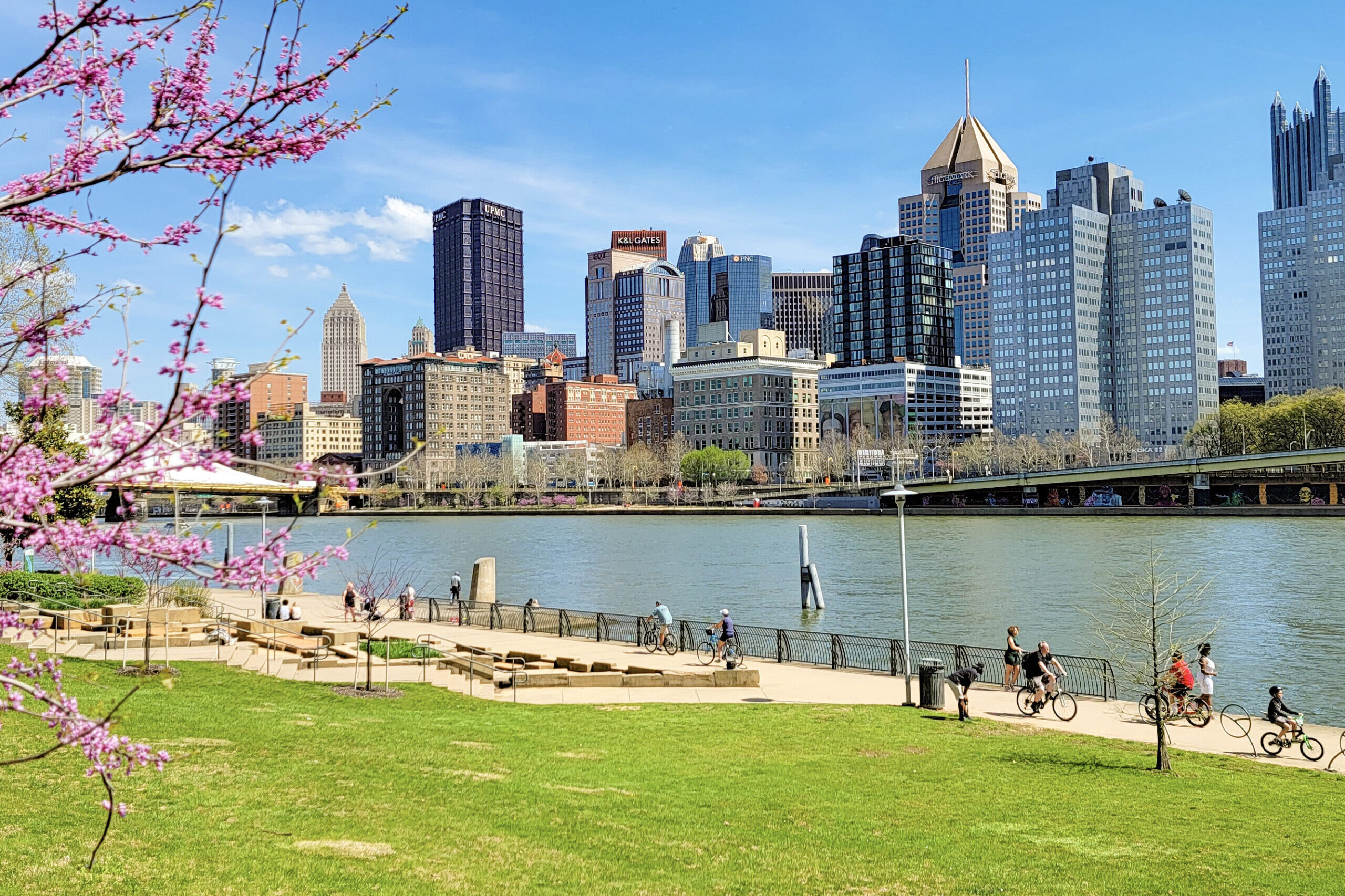 Bikers ride along a paved trail at the edge of a river with a view of Pittsburgh's city skyline in the background.