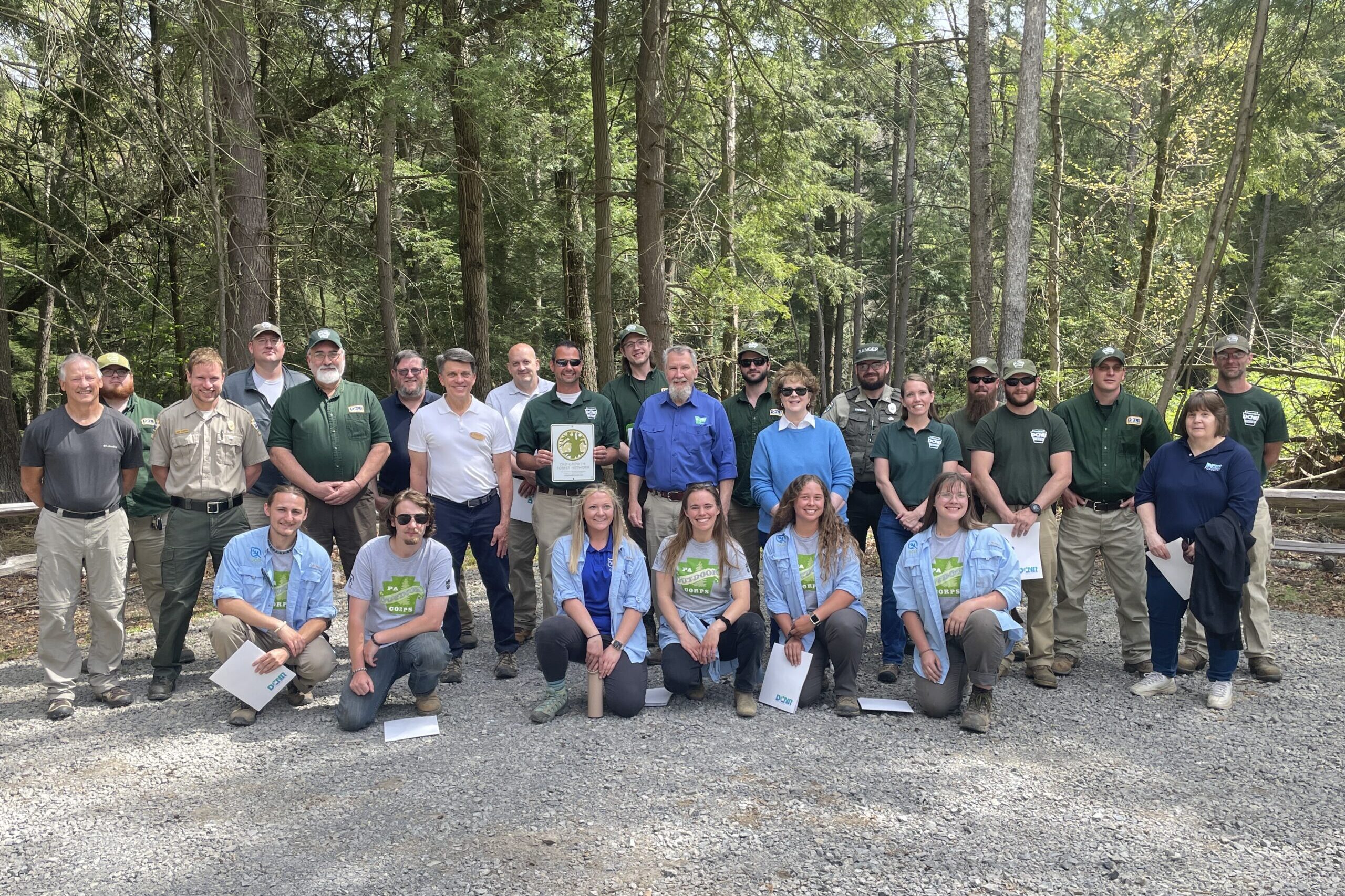 A big group stands together in front of the forest. One is holding their induction into the old growth certificate.