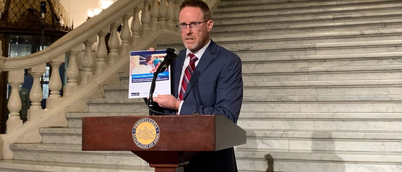 A man stands at a podium at the bottom of steps inside the state capitol holding a report.
