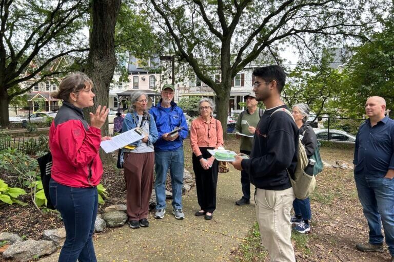 A woman with a clipboard stands under trees talking facing a man with a clipboard, in front of a group of people