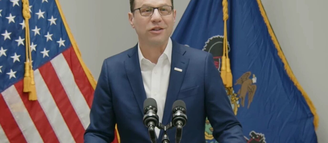 Governor Josh Shapiro stands behind a podium with the Pennsylvania and American flags behind them.