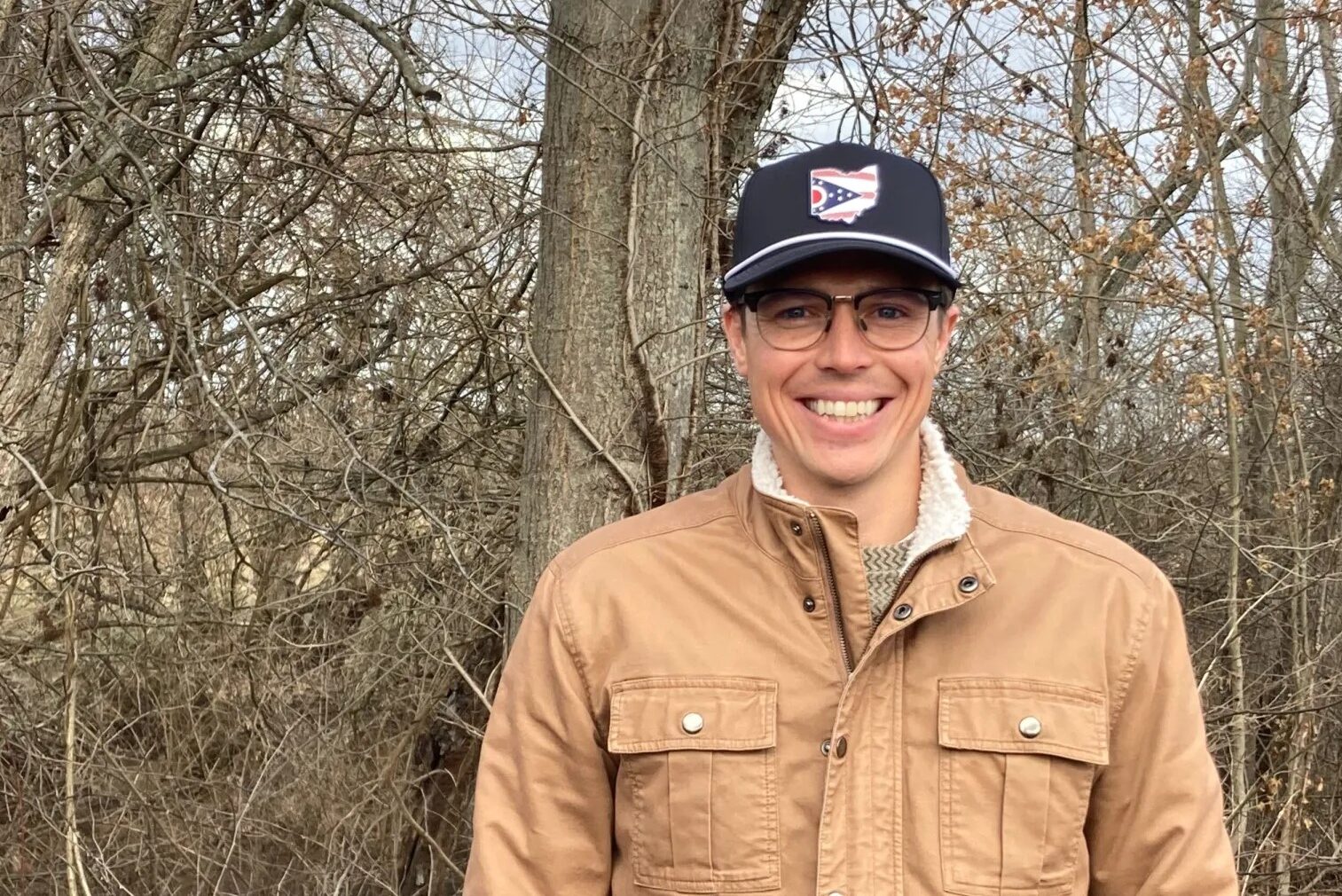 A man in a blue hat, glasses and brown jacket stands in front of a line of trees without leaves.