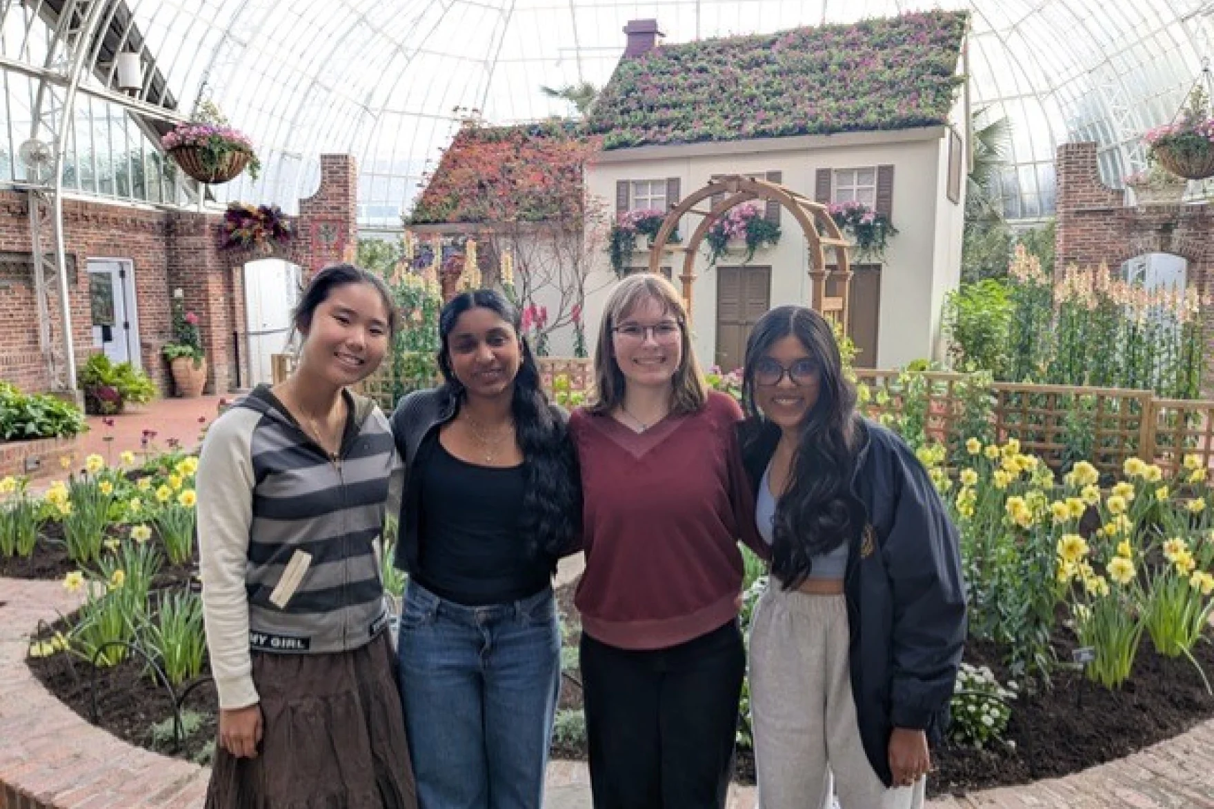 Four teenage girls stand in front of an exhibit of s cottage and flowers.