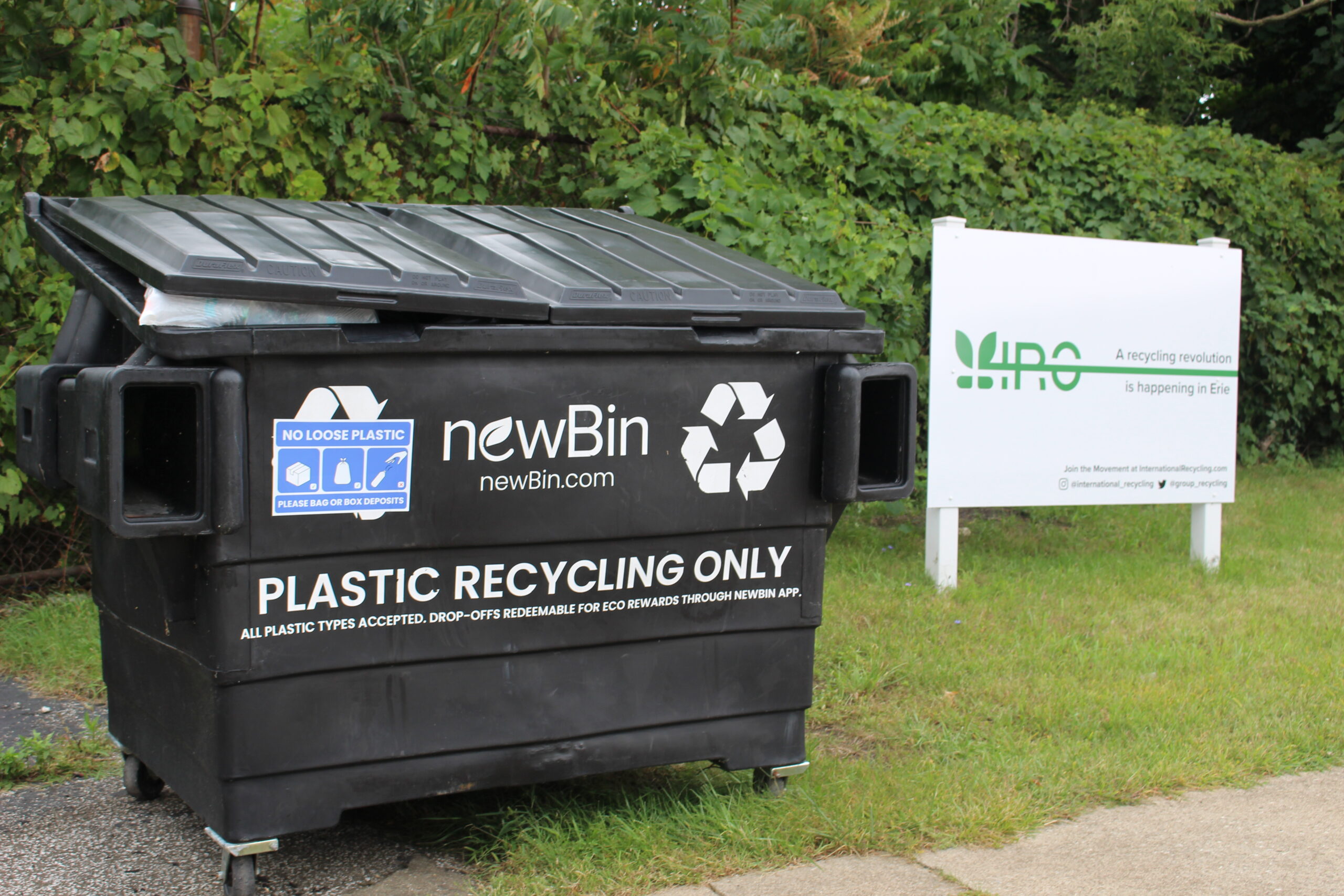 A dark colored community recycling bin, next to a white sign with the letter IRG in green