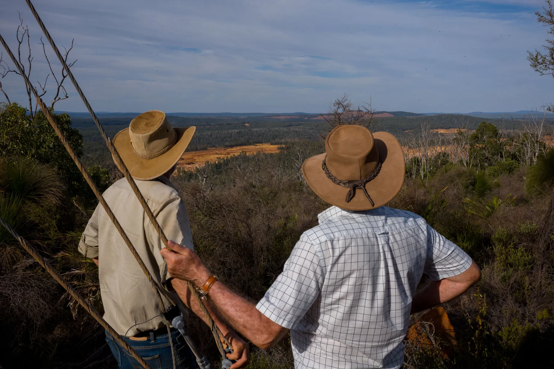 Two men with their backs to the camera,wearing cowboy hats ,look out over a mine in the distance.