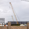 A crane looms above a construction site behind a wall, against a cloudy sky.